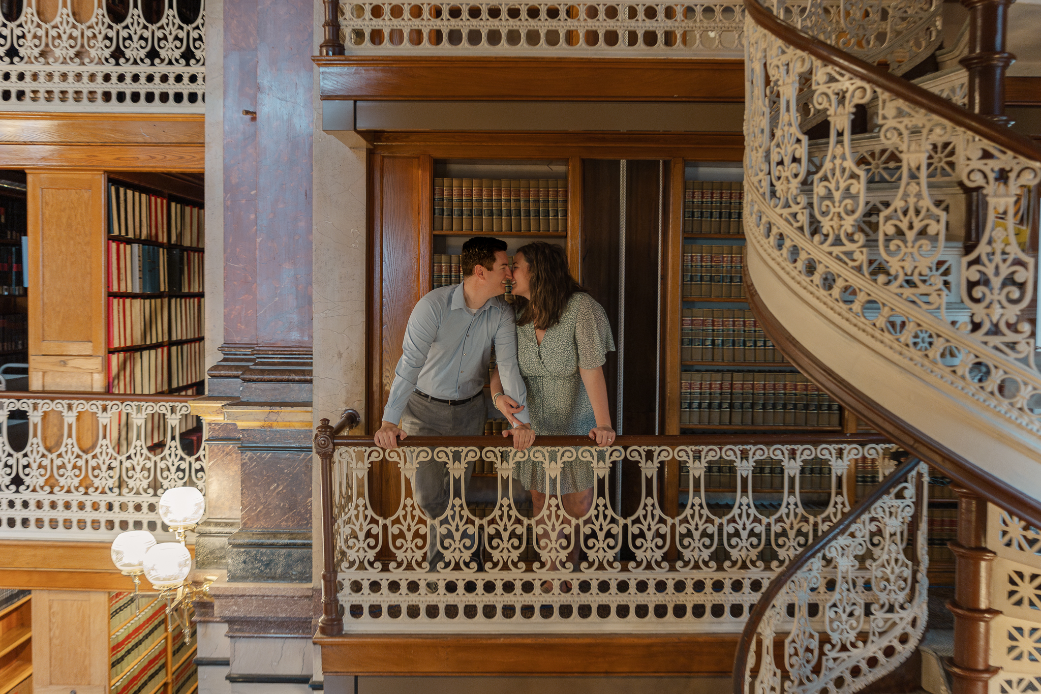 Couple leans in for a kiss on ornate balcony inside Iowa State Law Library during engagement session in Des Moines